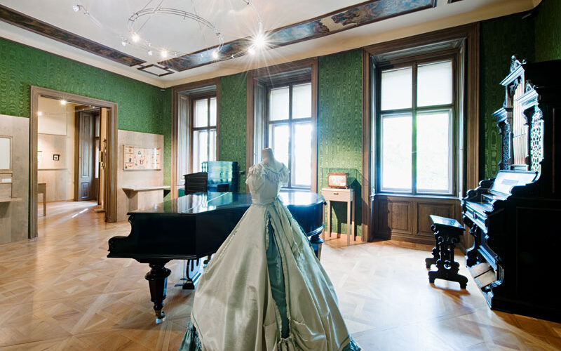 Eleganter historischer Raum mit grünen Tapeten, Holzböden, einem Flügel, einer Orgel und einem ausgestellten historischen Kleid. Große Fenster lassen viel natürliches Licht herein.