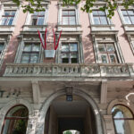 Ein beigefarbenes historisches Gebäude mit roten Fahnen, die über einem gewölbten Eingang hängen; zwei Personen gehen am Eingang vorbei, und am oberen Rand des Bildes sind grüne Baumzweige zu sehen.