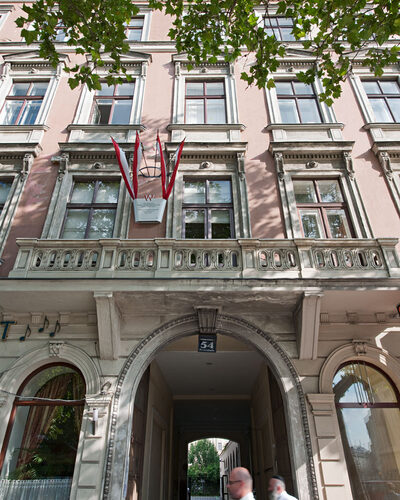 Ein beigefarbenes historisches Gebäude mit roten Fahnen, die über einem gewölbten Eingang hängen; zwei Personen gehen am Eingang vorbei, und am oberen Rand des Bildes sind grüne Baumzweige zu sehen.