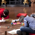 Die Kinder sitzen auf dem Boden und zeichnen und malen in Skizzenbüchern, während um sie herum in einem großen Innenraum Kunstmaterialien verstreut sind.