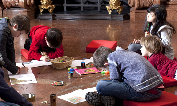 Die Kinder sitzen auf dem Boden und zeichnen und malen in Skizzenbüchern, während um sie herum in einem großen Innenraum Kunstmaterialien verstreut sind.