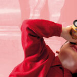 Ein Kind in einem roten Pullover schaut durch ein Kaleidoskop, während es sich vor einem rosafarbenen Hintergrund hinlegt.