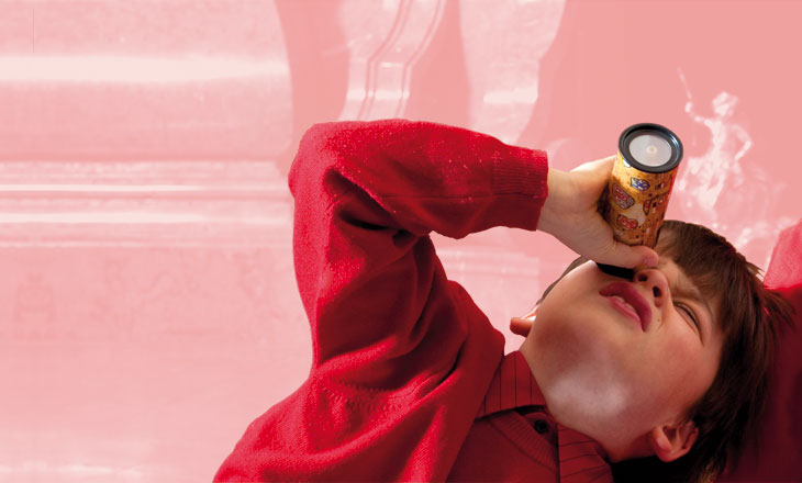 Ein Kind in einem roten Pullover schaut durch ein Kaleidoskop, während es sich vor einem rosafarbenen Hintergrund hinlegt.