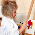 Ein kleines Kind malt mit roter und gelber Farbe auf einer Leinwand, steht an einer Staffelei und trägt ein weißes Hemd.