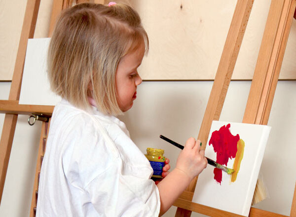 Ein kleines Kind malt mit roter und gelber Farbe auf einer Leinwand, steht an einer Staffelei und trägt ein weißes Hemd.