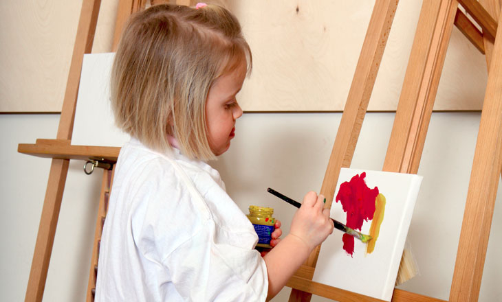 Ein kleines Kind malt mit roter und gelber Farbe auf einer Leinwand, steht an einer Staffelei und trägt ein weißes Hemd.