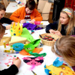Fünf Kinder sitzen um einen Tisch herum und basteln und verzieren Papier mit Markern, Kleber und verschiedenen bunten Materialien.