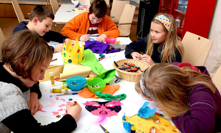 Fünf Kinder sitzen um einen Tisch herum und basteln und verzieren Papier mit Markern, Kleber und verschiedenen bunten Materialien.
