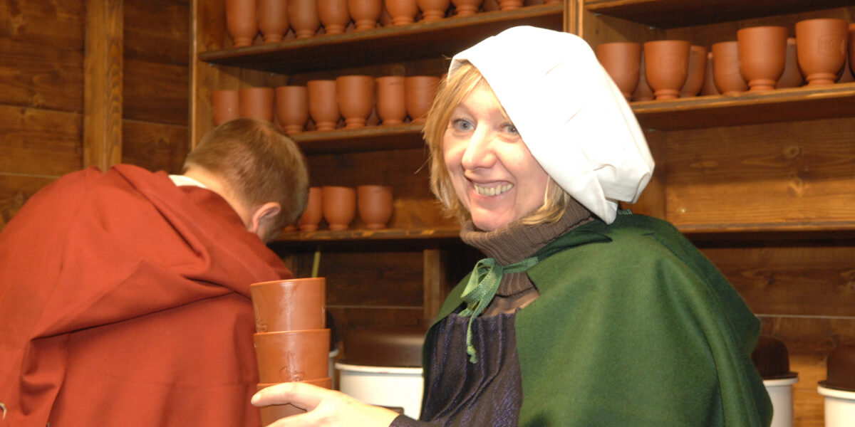 Eine Frau in historischer Kleidung hält gestapelte Tonbecher und lächelt, während ein Mann in ähnlicher Kleidung den Blick abwendet. Regale mit Tonbechern säumen die Holzwand hinter ihnen.