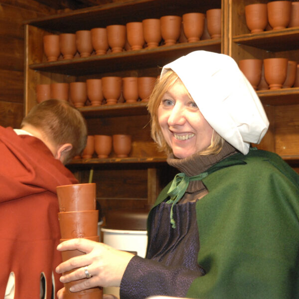 Eine Frau in historischer Kleidung hält gestapelte Tonbecher und lächelt, während ein Mann in ähnlicher Kleidung den Blick abwendet. Regale mit Tonbechern säumen die Holzwand hinter ihnen.