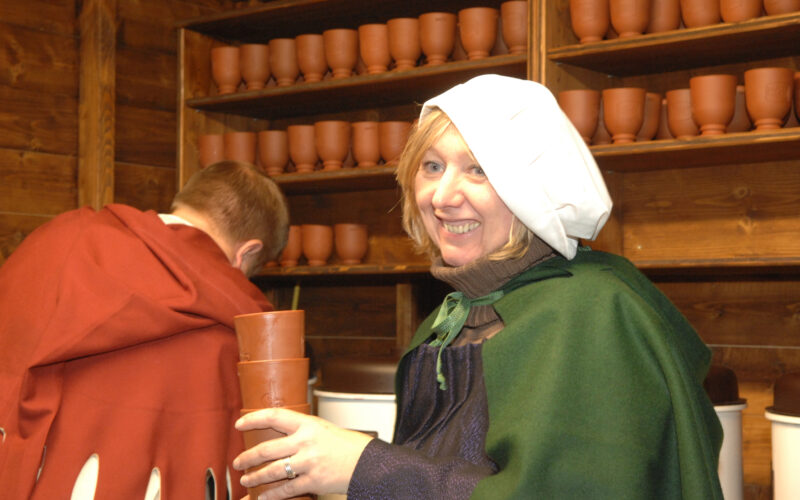 Eine Frau in historischer Kleidung hält gestapelte Tonbecher und lächelt, während ein Mann in ähnlicher Kleidung den Blick abwendet. Regale mit Tonbechern säumen die Holzwand hinter ihnen.