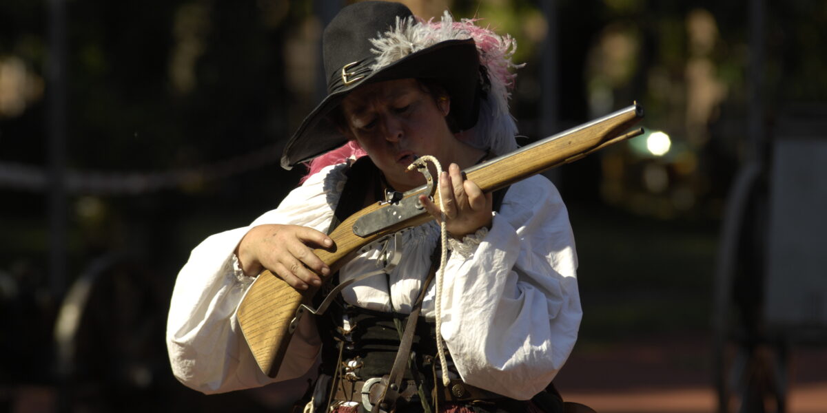 Eine Person in historischem Kostüm hält und untersucht eine Muskete im Freien, mit Sonnenlicht und verschwommenem Grün im Hintergrund.