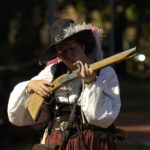 Eine Person in historischem Kostüm hält und untersucht eine Muskete im Freien, mit Sonnenlicht und verschwommenem Grün im Hintergrund.