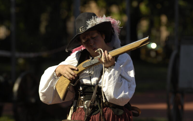 Eine Person in historischem Kostüm hält und untersucht eine Muskete im Freien, mit Sonnenlicht und verschwommenem Grün im Hintergrund.