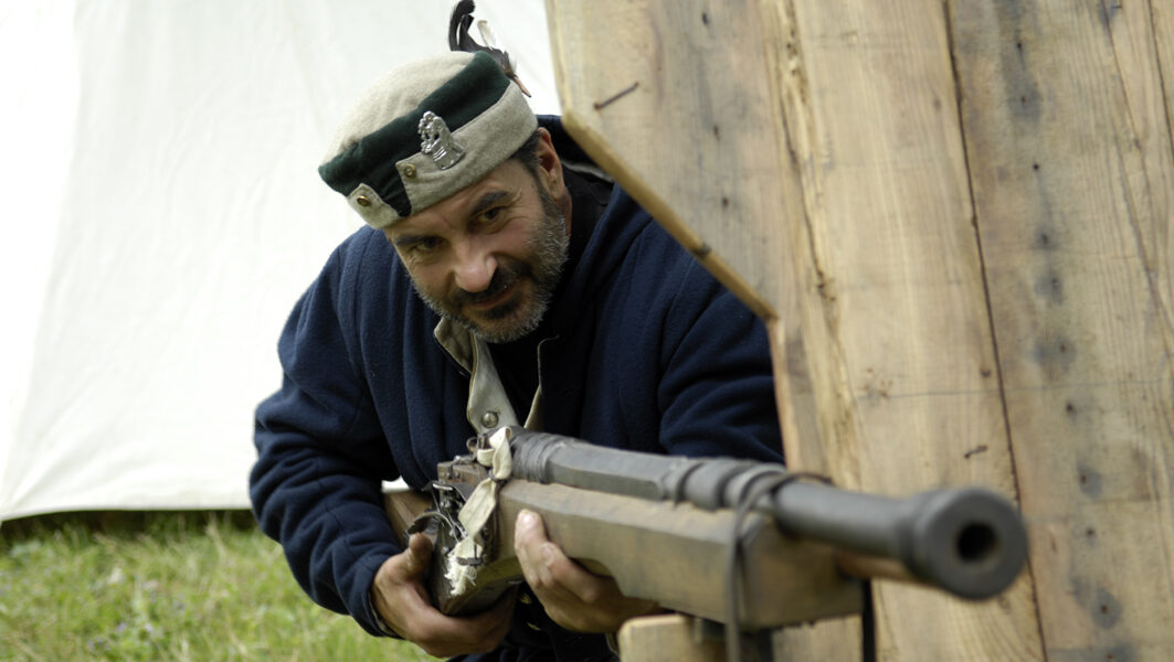 Mann in historischer Kleidung zielt mit einer Muskete durch eine hölzerne Barriere, im Hintergrund ein weißes Zelt.
