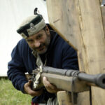 Mann in historischer Kleidung zielt mit einer Muskete durch eine hölzerne Barriere, im Hintergrund ein weißes Zelt.