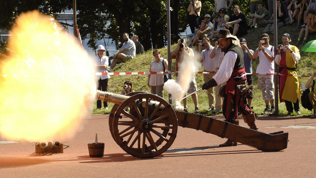 Ein Mann in einem historischen Kostüm feuert im Freien eine Kanone ab, während eine Menschenmenge hinter einer Sicherheitsabsperrung zusieht und Fotos macht.