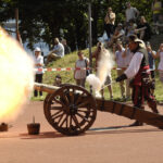 Ein Mann in einem historischen Kostüm feuert im Freien eine Kanone ab, während eine Menschenmenge hinter einer Sicherheitsabsperrung zusieht und Fotos macht.
