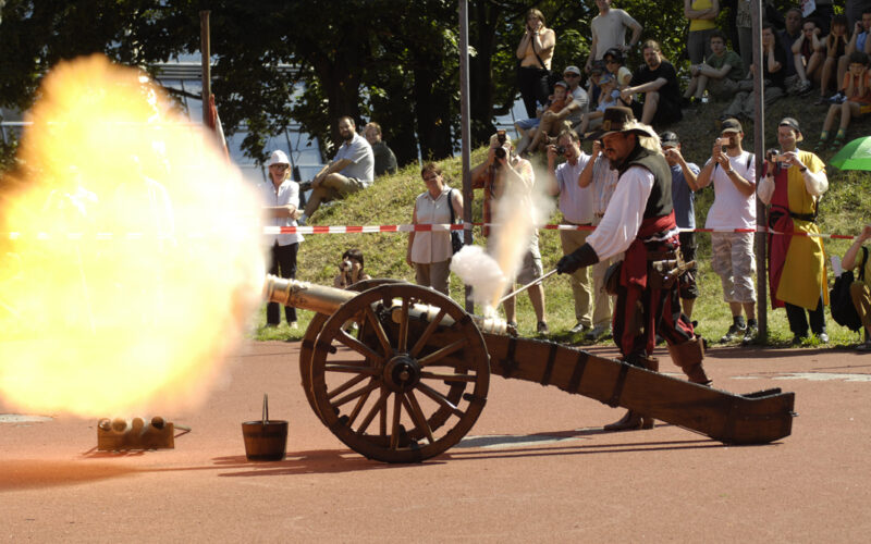 Ein Mann in einem historischen Kostüm feuert im Freien eine Kanone ab, während eine Menschenmenge hinter einer Sicherheitsabsperrung zusieht und Fotos macht.