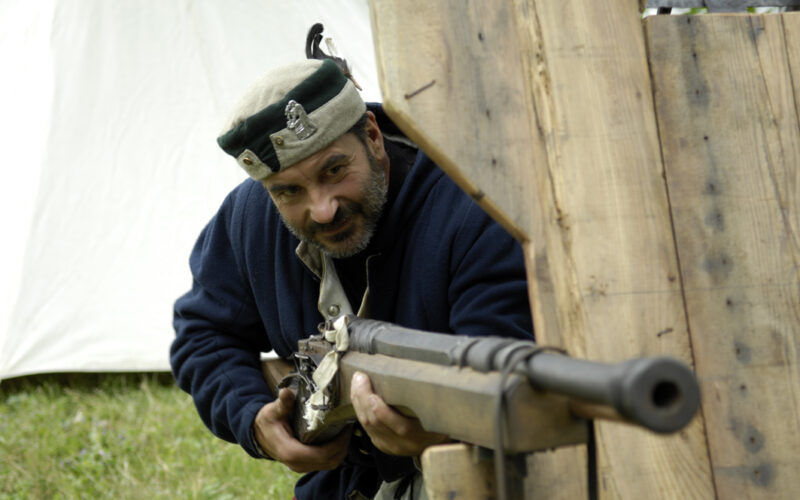 Ein Mann in historischer Kleidung zielt mit einer Muskete durch eine Lücke in einer Holzbarrikade im Freien.