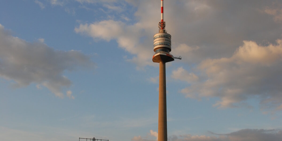Ein hoher Fernmeldeturm erhebt sich vor einem teilweise bewölkten Himmel über einer Menschenmenge, die sich bei einer Veranstaltung im Freien versammelt hat, mit sichtbaren Zelten der Marke NIVEA.
