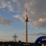 Ein hoher Fernmeldeturm erhebt sich vor einem teilweise bewölkten Himmel über einer Menschenmenge, die sich bei einer Veranstaltung im Freien versammelt hat, mit sichtbaren Zelten der Marke NIVEA.