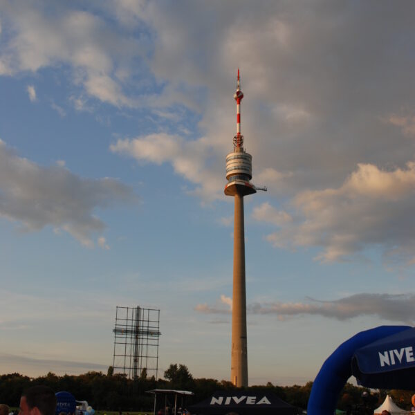 Ein hoher Fernmeldeturm erhebt sich vor einem teilweise bewölkten Himmel über einer Menschenmenge, die sich bei einer Veranstaltung im Freien versammelt hat, mit sichtbaren Zelten der Marke NIVEA.