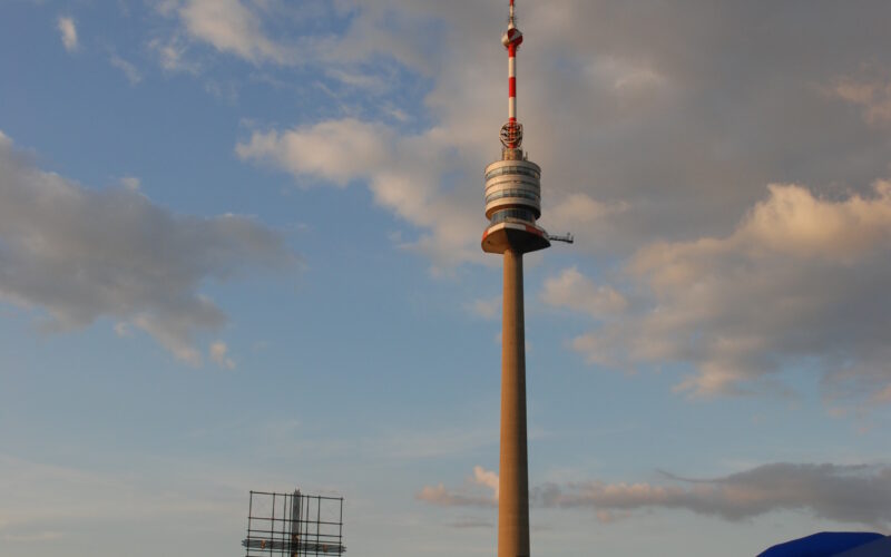 Ein hoher Fernmeldeturm erhebt sich vor einem teilweise bewölkten Himmel über einer Menschenmenge, die sich bei einer Veranstaltung im Freien versammelt hat, mit sichtbaren Zelten der Marke NIVEA.