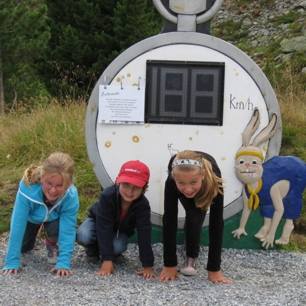 Drei Kinder kauern an einer Startlinie vor einer Stoppuhr, die auf einem Schild mit einem Cartoon-Hasenmotiv angebracht ist, im Freien auf einem Kiesweg.