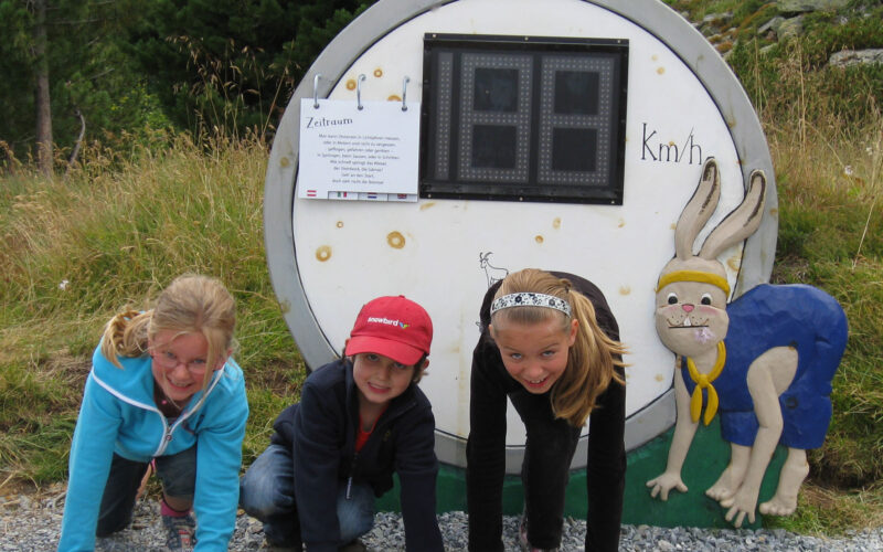 Drei Kinder kauern an einer Startlinie vor einer Stoppuhr, die auf einem Schild mit einem Cartoon-Hasenmotiv angebracht ist, im Freien auf einem Kiesweg.