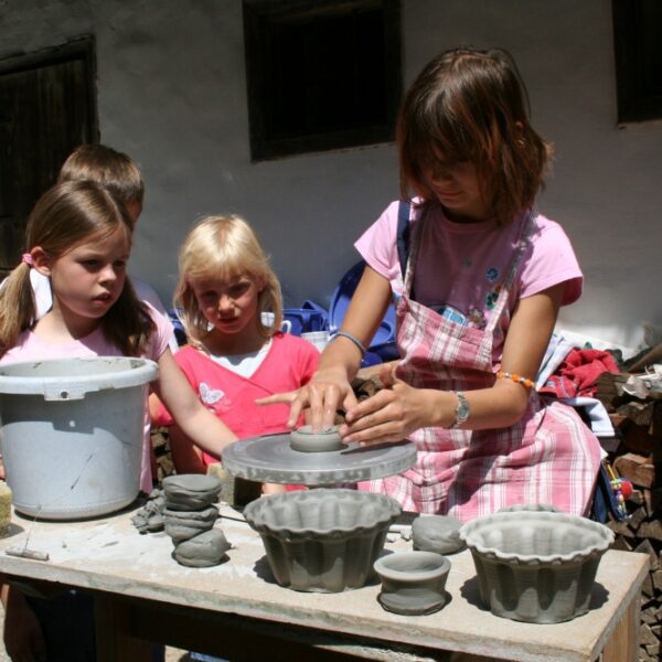 Eine Gruppe von Kindern steht im Freien um einen Tisch herum, während ein Mädchen auf einer Töpferscheibe Ton formt, während auf dem Tisch Tonobjekte und Formen ausgestellt sind.