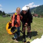 Ein Mädchen und ein Erwachsener mit Gleitschirmausrüstung stehen zusammen auf einer grasbewachsenen Wiese mit Bergen und Häusern im Hintergrund.