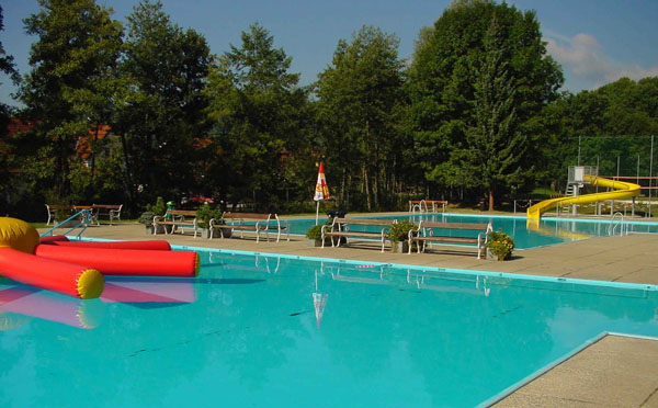 Freibad mit klarem, blauem Wasser, einem aufblasbaren Schwimmer, einer gelben Wasserrutsche, Bänken und Bäumen im Hintergrund an einem sonnigen Tag.