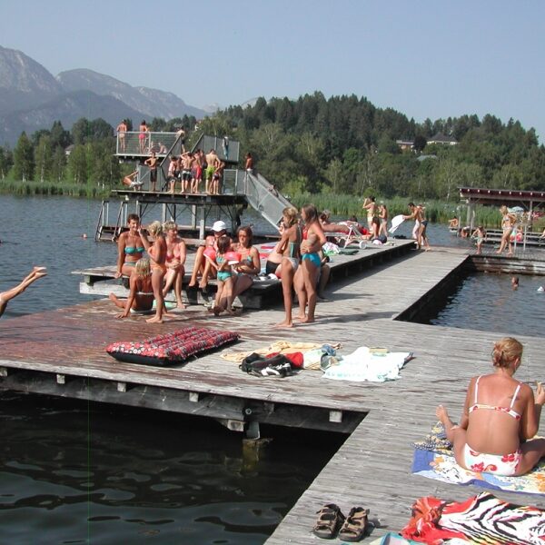 Die Menschen sonnen sich und schwimmen auf hölzernen Stegen an einem See mit Sprungtürmen, umgeben von Bäumen und Bergen im Hintergrund.