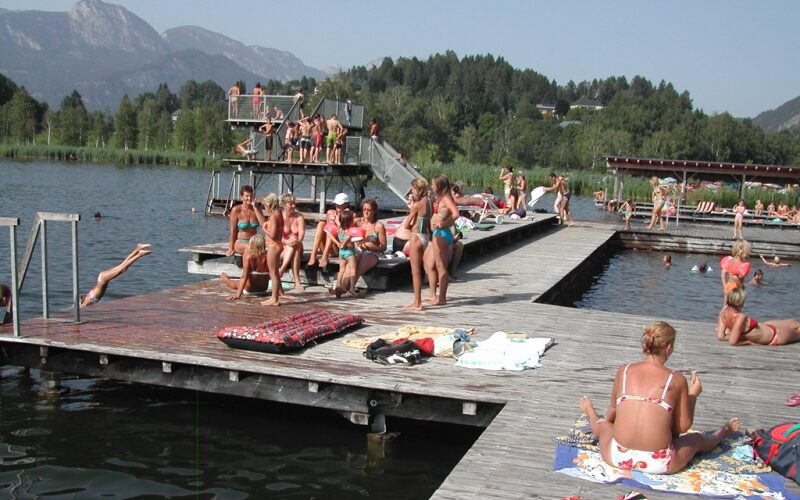 Die Menschen sonnen sich und schwimmen auf hölzernen Stegen an einem See mit Sprungtürmen, umgeben von Bäumen und Bergen im Hintergrund.