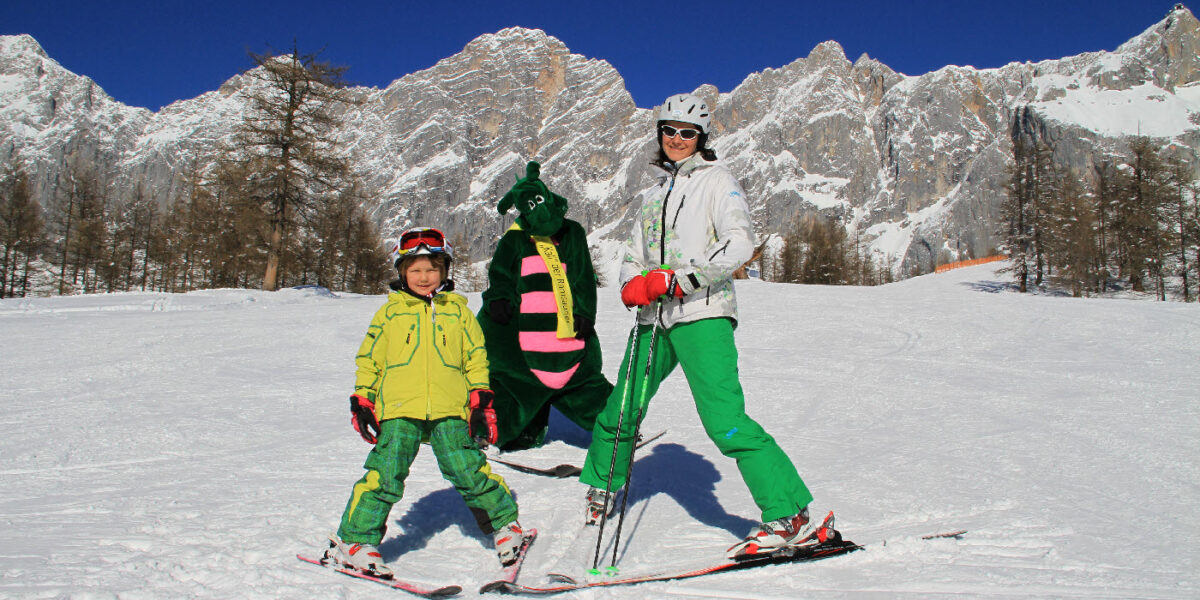 Ein Erwachsener und ein Kind in grüner Skiausrüstung posieren auf einer verschneiten Piste mit einer Person in einem grünen Drachenkostüm, im Hintergrund sind Berge und Bäume zu sehen.