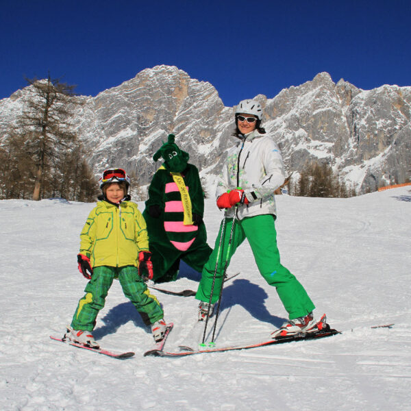 Ein Erwachsener und ein Kind in grüner Skiausrüstung posieren auf einer verschneiten Piste mit einer Person in einem grünen Drachenkostüm, im Hintergrund sind Berge und Bäume zu sehen.