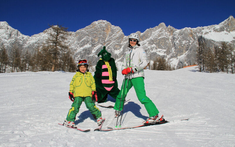 Ein Erwachsener und ein Kind in grüner Skiausrüstung posieren auf einer verschneiten Piste mit einer Person in einem grünen Drachenkostüm, im Hintergrund sind Berge und Bäume zu sehen.