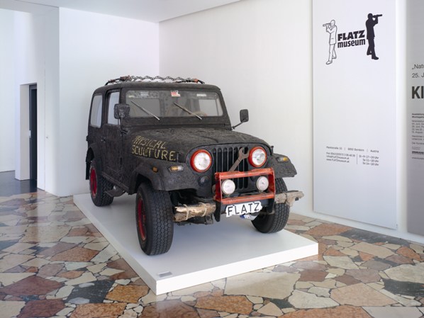 Ein schwarzer, verwitterter Jeep mit roten Rädern steht auf einer weißen Plattform in einem Museum, mit dem Nummernschild FLATZ" und der Aufschrift MUSICAL SCULPTURE" auf der Motorhaube.