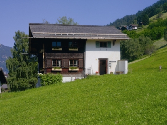 Ein zweistöckiges Haus mit hölzernen oberen Wänden und weißen unteren Wänden steht an einem grasbewachsenen Hang, umgeben von Bäumen und Hügeln unter einem klaren blauen Himmel.