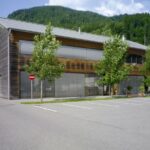 Ein modernes zweistöckiges Holzgebäude mit großen Fenstern, umgeben von Bäumen und Bergen, angrenzend an einen leeren Parkplatz und ein "Betreten verboten"-Schild.