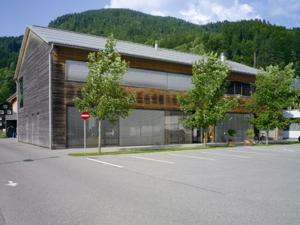 Ein modernes zweistöckiges Holzgebäude mit großen Fenstern, umgeben von Bäumen und Bergen, angrenzend an einen leeren Parkplatz und ein "Betreten verboten"-Schild.