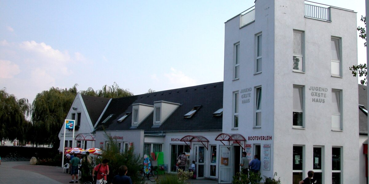 Ein weißes, modernes Gästehaus mit eckigen Dächern und großen Fenstern; draußen stehen Menschen mit Fahrrädern, und im Erdgeschoss sind ein paar Geschäfte zu sehen.