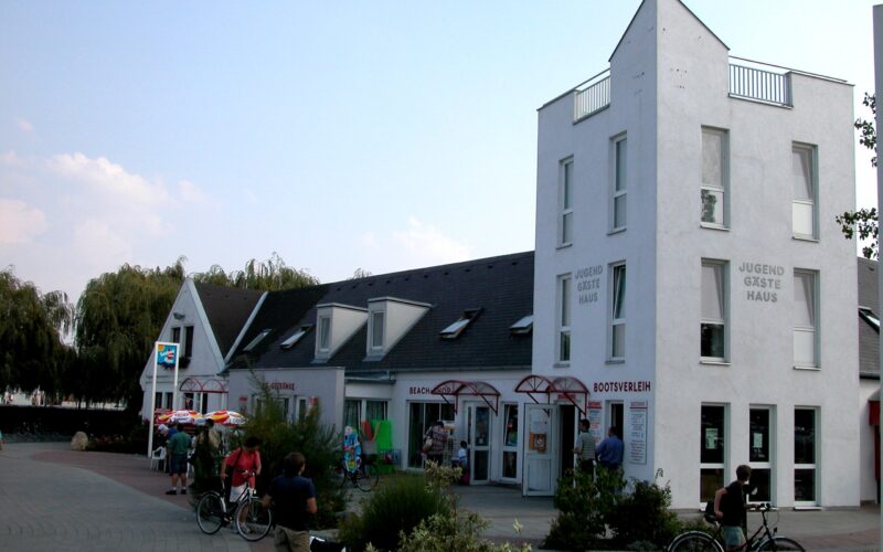 Ein weißes, modernes Gästehaus mit eckigen Dächern und großen Fenstern; draußen stehen Menschen mit Fahrrädern, und im Erdgeschoss sind ein paar Geschäfte zu sehen.