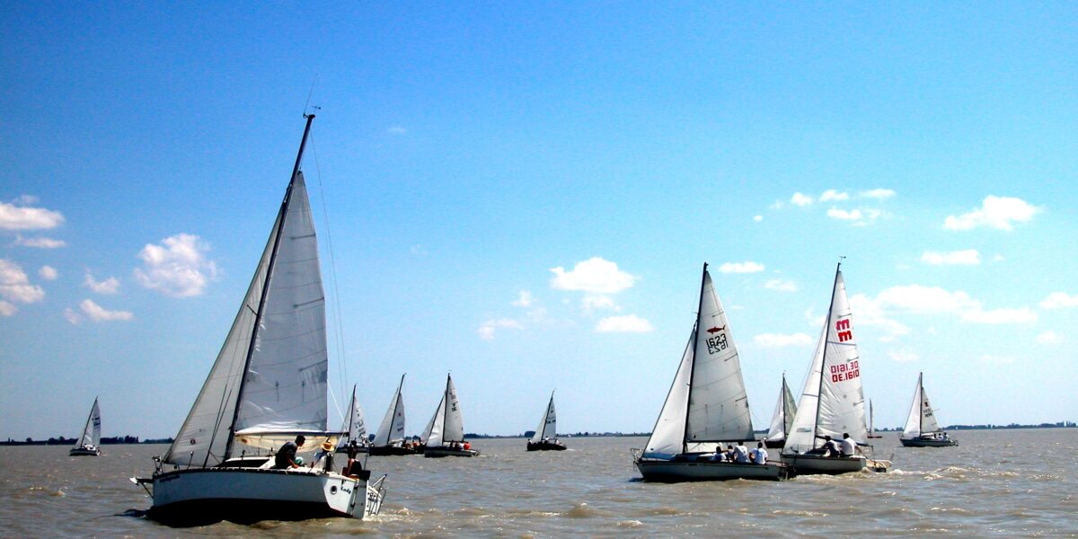 Mehrere Segelboote mit weißen Segeln liegen auf einer großen Wasserfläche unter einem klaren blauen Himmel mit vereinzelten Wolken.