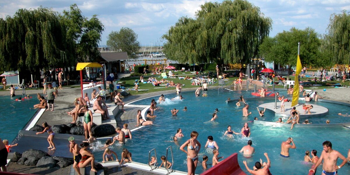 Öffentliches Freibad mit vielen Menschen, die an einem sonnigen Tag schwimmen, spielen und sich entspannen; Kinder benutzen eine rote Wasserrutsche im Vordergrund und eine Grasfläche im Hintergrund.
