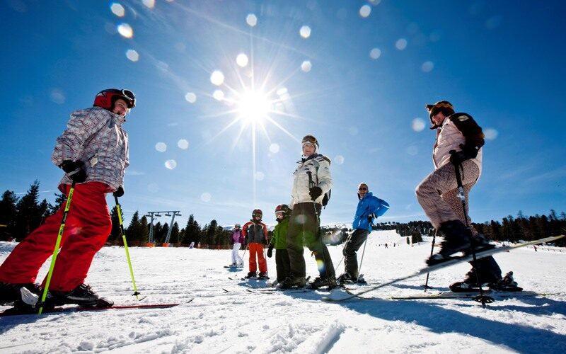 Eine Gruppe von Menschen steht in Winterkleidung und mit Skistöcken auf einer verschneiten Skipiste im hellen Sonnenlicht, über der ein strahlend blauer Himmel liegt.