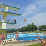 Öffentliches Freibad mit einem hohen Sprungturm; mehrere Personen schwimmen, sonnen sich, und eine Person springt in der Luft ins Wasser. Bäume und Sonnenschirme sind im Hintergrund zu sehen.