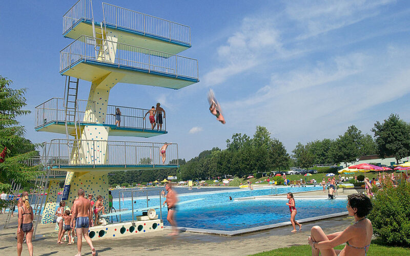 Öffentliches Freibad mit einem hohen Sprungturm; mehrere Personen schwimmen, sonnen sich, und eine Person springt in der Luft ins Wasser. Bäume und Sonnenschirme sind im Hintergrund zu sehen.