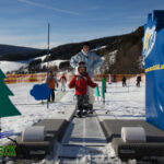 Ein Kind mit Helm steht in Begleitung eines Erwachsenen an einem Förderband-Skilift in einem verschneiten Skigebiet mit Sicherheitsbarrieren und dekorativen Aussparungen.